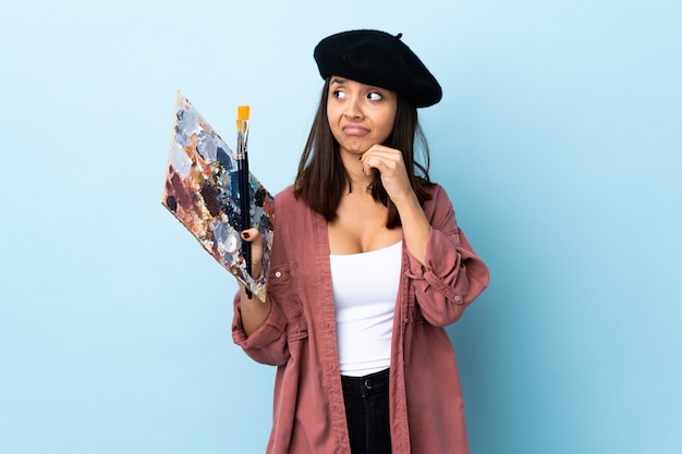 Foto mulher jovem artista segurando uma paleta sobre azul isolado tendo dúvidas e pensando.