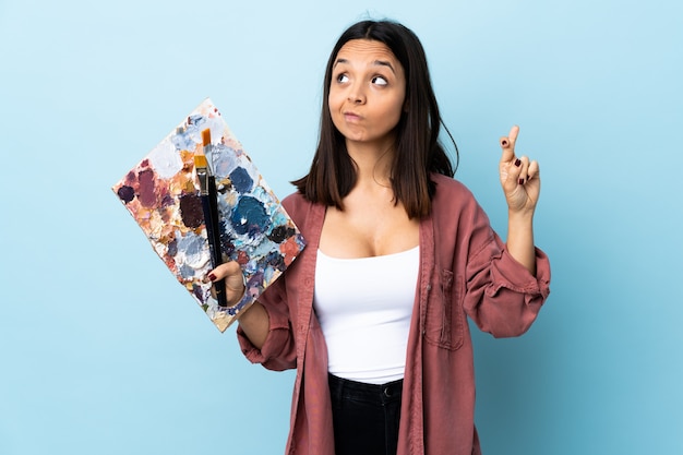 Mulher jovem artista segurando uma paleta sobre parede azul com dedos cruzando e desejando o melhor