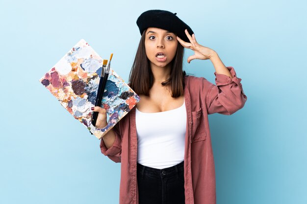 Mulher jovem artista segurando uma paleta sobre parede azul com expressão de surpresa