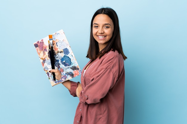 Mulher jovem artista segurando uma paleta sobre parede azul com os braços cruzados e olhando para a frente.