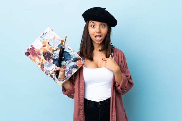 Mulher jovem artista segurando uma paleta sobre parede azul isolada com expressão facial de surpresa