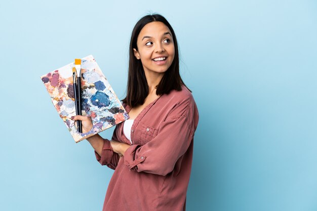 Mulher jovem artista segurando uma paleta sobre parede azul isolada com os braços cruzados e feliz