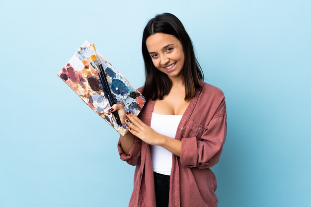 Mulher jovem artista segurando uma paleta sobre parede azul mantém a palma da mão unida