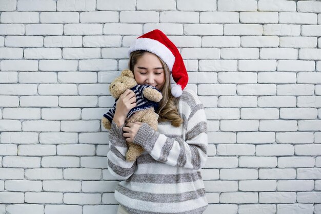 Mulher jovem asiática bonita do retrato no traje do natal e usar chapéu de papai noel vermelho e segurar um ursinho de pelúcia com sorriso no fundo de tijolos brancos, copie o espaço. feliz por uma mulher adolescente no tema natal.