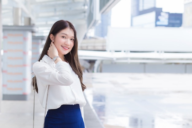 Mulher jovem asiática confiante que trabalha, tem cabelo comprido e uma camisa branca, está em pé ao ar livre urbano enquanto caminha para o escritório em uma cidade grande com prédios comerciais com a cidade como pano de fundo