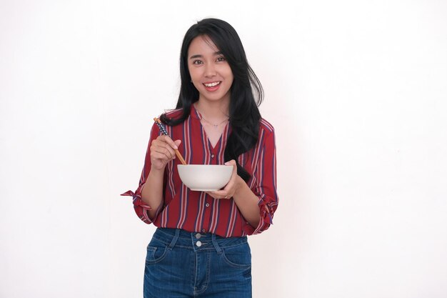 Mulher jovem asiática de pé e comendo sua comida de uma tigela branca usando pauzinhos