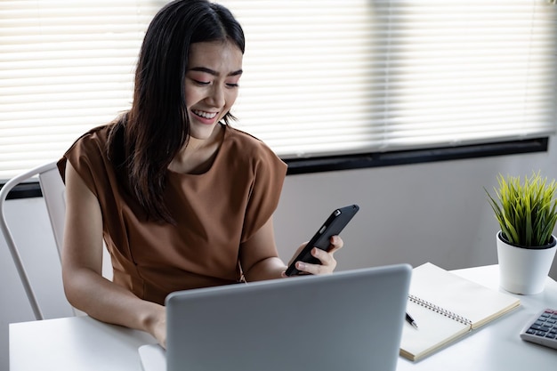 Mulher jovem asiática jogando social online no celular com laptop está na mesa por um sorriso feliz na sala de trabalho em casa. Mulher feliz jogando internet online no smartphone.