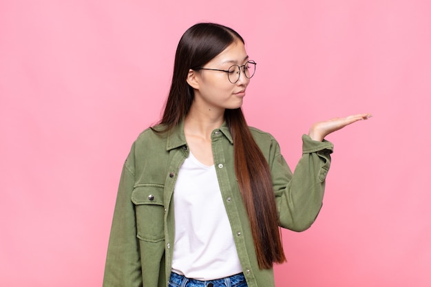 Mulher jovem asiática se sentindo feliz e sorrindo casualmente, olhando para um objeto ou conceito seguro ao lado