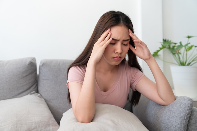 Mulher jovem asiática segurando sua cabeça, ela tem uma dor de cabeça sentada no sofá-sofá na sala de estar em casa.