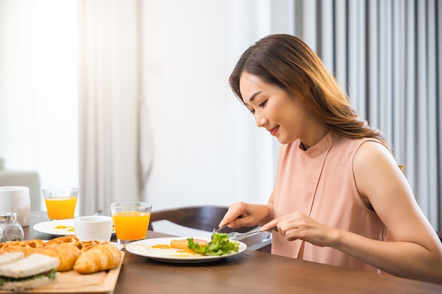 Mulher jovem asiática sentada na mesa da cozinha tomando café da manhã saudável em casa, mulher comendo café da manhã fresco servido com bebida antes de ir trabalhar de manhã em casa