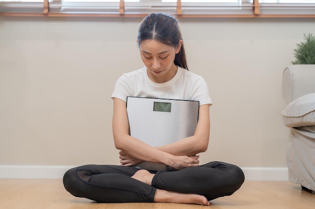 Mulher jovem asiática triste deprimida e estressada cobre o rosto com a mão sentada no chão pesando na balança na frente dela ganhar perder peso e gordura com excesso de peso pessoas dieta insalubre conceito infeliz