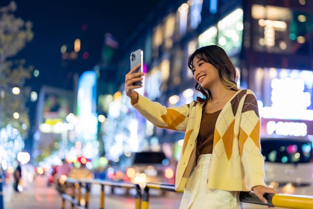 Mulher jovem asiática usando telefone móvel tirando selfie com paisagem urbana e luzes de rua à noite Menina atraente desfrutar e diversão vida noturna ao ar livre viagem cidade com o uso de tecnologia de dispositivo portátil