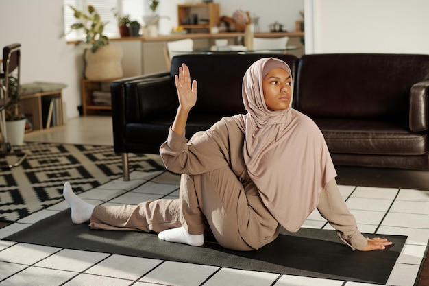 Mulher jovem ativa de hijab e macacão sentada no tapete no chão da sala de estar