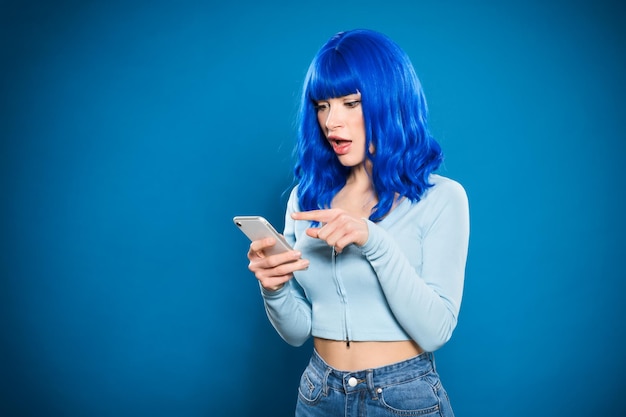 Mulher jovem atônita com cabelo azul olhando para a tela do celular e apontando o dedo em pé contra um fundo iluminado vívido