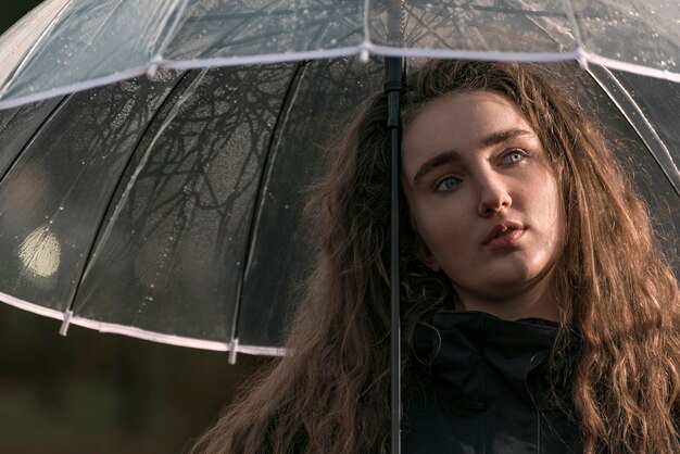 Mulher jovem atraente e pensativa com cabelos castanhos exuberantes sob guarda-chuva transparente Retrato de outono Imagem triste