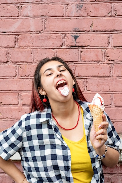 Foto mulher jovem atraente em roupas de verão comendo sorvete no fundo da parede de tijolos rosa na rua