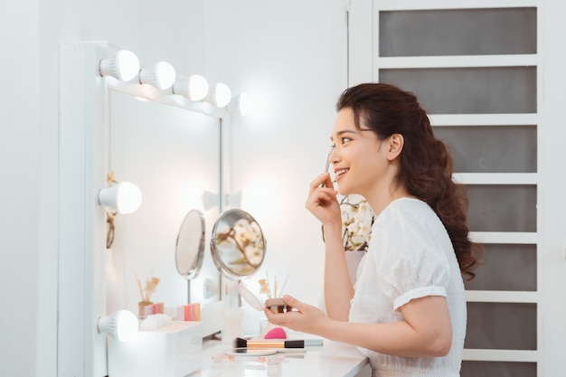 Mulher jovem atraente feliz aplicando cosméticos no rosto perto da mesa de cosméticos de manhã