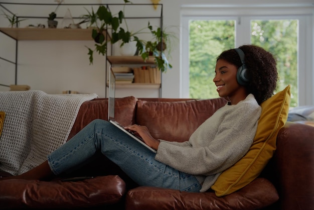 Mulher jovem atraente usando um laptop enquanto ouve música em fones de ouvido e relaxa em um sofá em casa