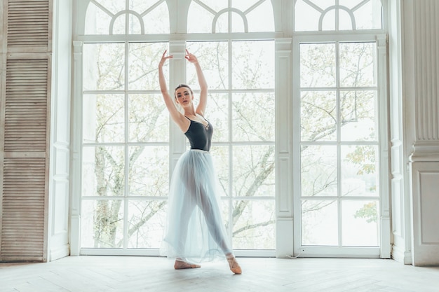 Mulher jovem bailarina clássica na aula de dança. A bailarina graciosa bonita pratica posições de balé na saia azul do tutu perto da janela grande no salão da luz branca.
