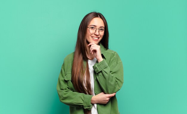 Mulher jovem, bem casual, parecendo feliz e sorrindo com a mão no queixo, pensando ou fazendo uma pergunta, comparando opções