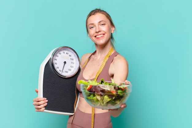 Mulher jovem bonita com expressão feliz e segurando uma balança e uma salada