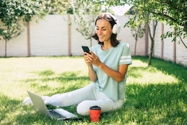Mulher jovem bonita feliz confiante moderna em fones de ouvido com um smartphone está trabalhando em um laptop enquanto está sentado no gramado.