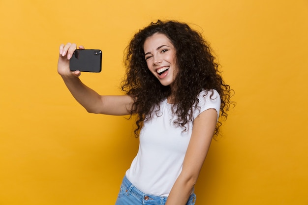 Mulher jovem bonita feliz posando isolada em amarelo tome uma selfie pelo telefone móvel.