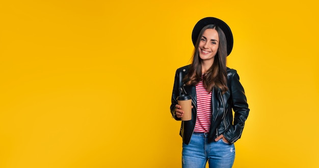 Mulher jovem bonita hippie sorridente com uma jaqueta preta e um chapéu com uma xícara de café ou chá na mão enquanto ela posa isolada em um fundo amarelo