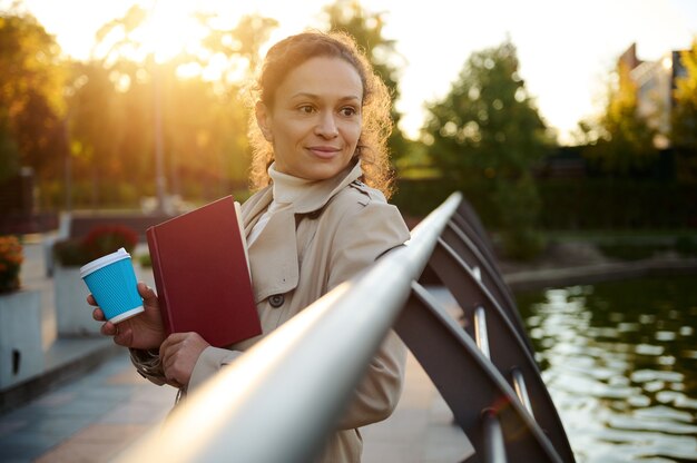 Mulher jovem bonita repousa em um parque outonal da cidade, segurando uma caneca de papel para viagem de café e um livro de mão, sorri, olhando para a câmera com raios de sol caindo no fundo das folhas amarelas douradas. Outono