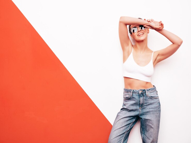 Foto mulher jovem, bonita, sorridente, de moda, de verão, camiseta branca e calças de ganga azuis, mulher sexy e despreocupada posando perto da parede no interior do estúdio, modelo positivo, com óculos de sol, alegre e feliz.
