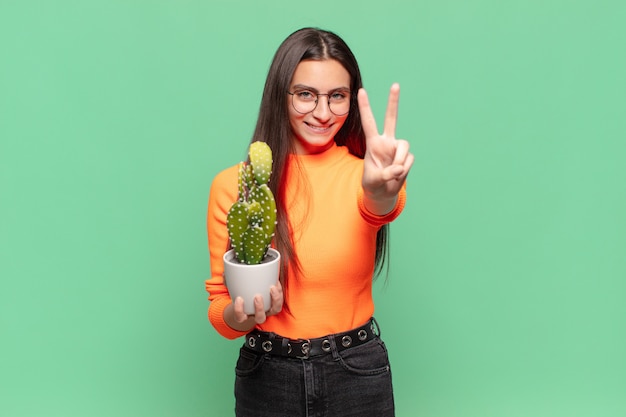 Mulher jovem bonita sorrindo e parecendo feliz, despreocupada e positiva, gesticulando vitória ou paz com uma mão. conceito de cacto