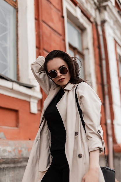 Mulher jovem bonita urbana elegante na moda primavera jovem sobretudo em óculos de sol da moda fica perto de uma parede vintage na rua. Hipster de linda garota posando ao ar livre na cidade. Look casual.