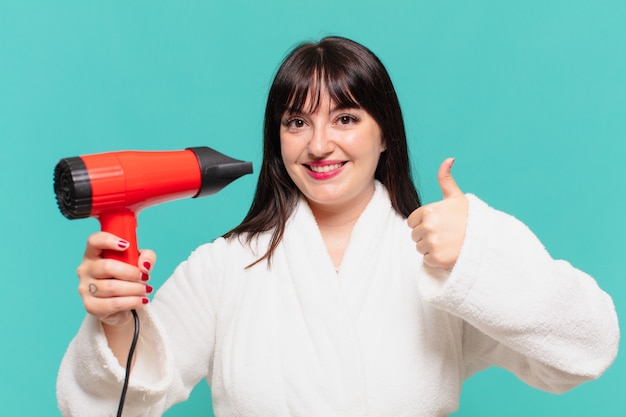 Mulher jovem bonita vestindo roupão de banho expressão feliz e secador de cabelo