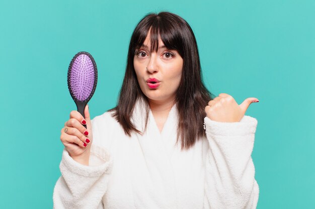Mulher jovem bonita vestindo um roupão de banho com expressão de surpresa e segurando uma escova de cabelo