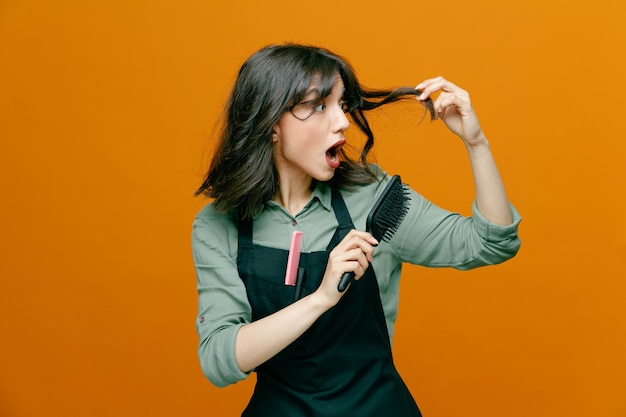 Mulher jovem cabeleireira vestindo avental segurando escova de cabelo penteando o cabelo parecendo preocupado em pé sobre fundo laranja