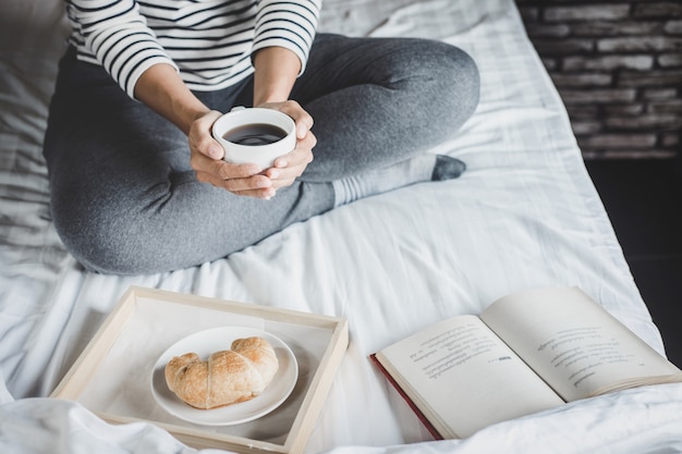 Mulher jovem, cama, com, livro velho, e, xícara café manhã, em, mãos, e, croissant