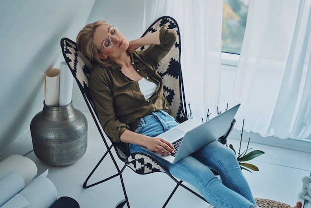 Mulher jovem cansada carregando laptop e mantendo os olhos fechados enquanto está sentada em uma cadeira confortável em casa