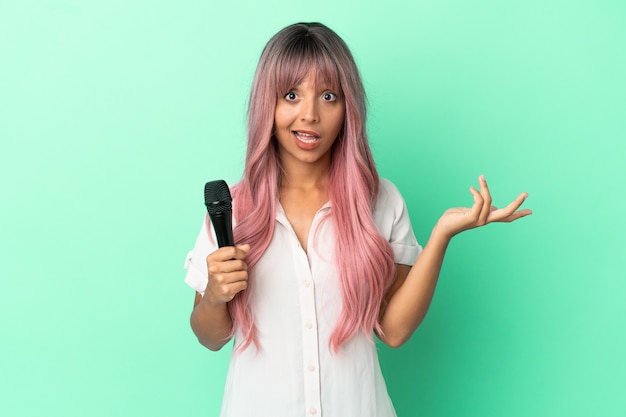 Mulher jovem cantora de raça mista com cabelo rosa isolado em um fundo verde com expressão facial chocada