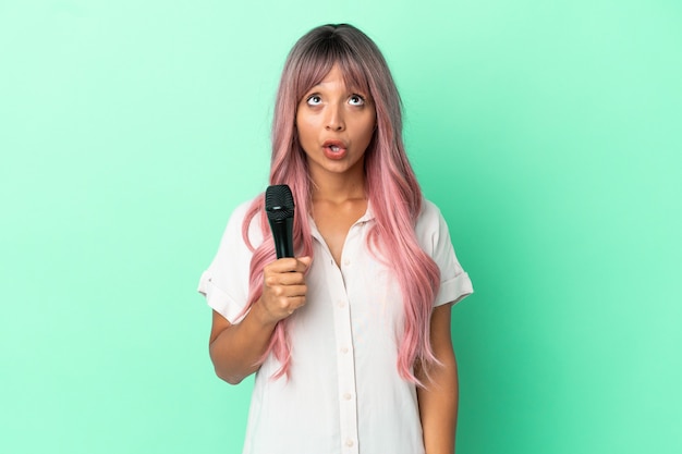 Mulher jovem cantora de raça mista com cabelo rosa isolado em um fundo verde, olhando para cima e com expressão de surpresa