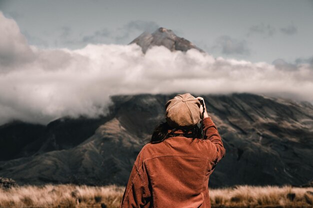 Foto mulher jovem caucasiana da parte de trás vestindo um casaco castanho fotografando a montanha grande desfocada e