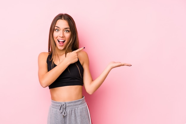 Mulher jovem caucasiano fitness posando em uma parede rosa animado segurando um espaço de cópia na palma da mão.