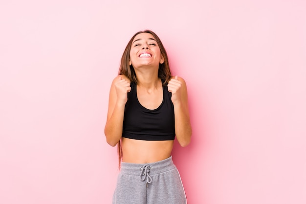 Mulher jovem caucasiano fitness posando em uma parede rosa, levantando o punho, sentindo-se feliz e bem sucedido. conceito de vitória