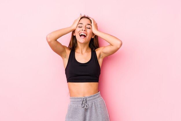 Mulher jovem caucasiano fitness posando em uma parede rosa ri alegremente, mantendo as mãos na cabeça.