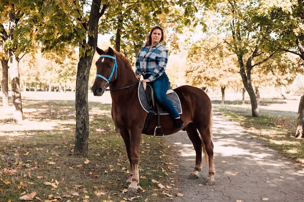 Mulher jovem, cavalgando um cavalo marrom, caminhando no parque de outono em um dia ensolarado