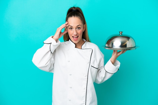 Mulher jovem chef caucasiano com bandeja isolada em fundo azul com expressão de surpresa