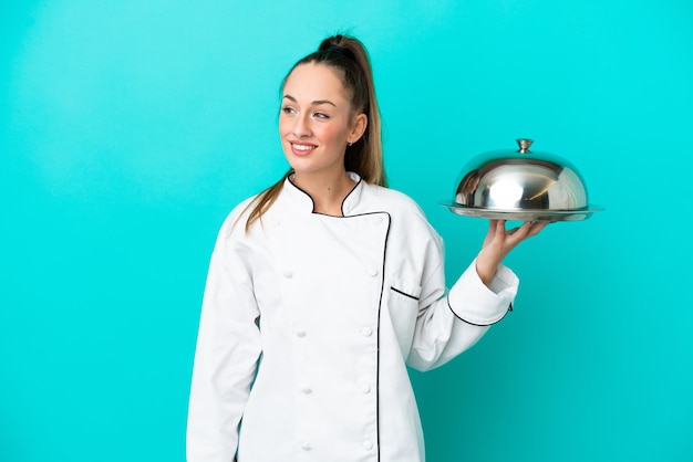 Mulher jovem chef caucasiano com bandeja isolada em fundo azul, olhando para o lado e sorrindo