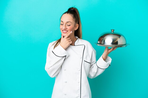 Mulher jovem chef caucasiano com bandeja isolada em fundo azul, olhando para o lado e sorrindo