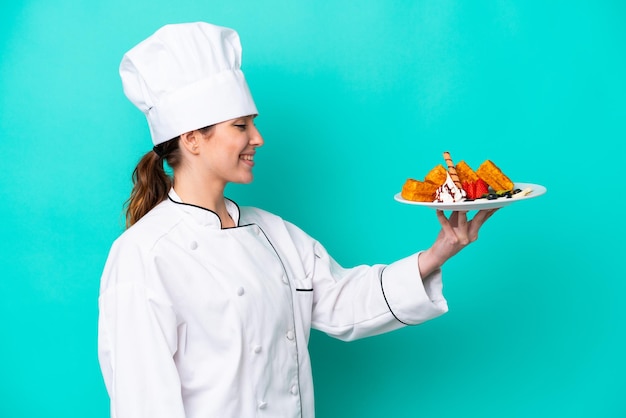 Mulher jovem chef caucasiano segurando waffles isolados em fundo azul com expressão feliz