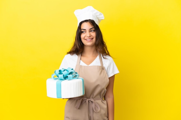 Mulher jovem chef de confeitaria caucasiana com um grande bolo isolado em um fundo amarelo pensando uma ideia enquanto olha para cima