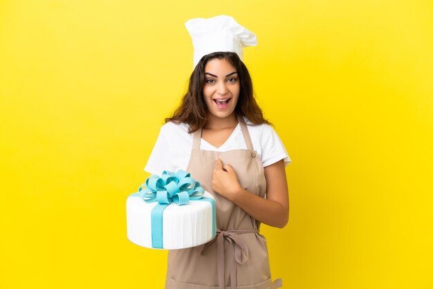 Mulher jovem chef de pastelaria caucasiana com um grande bolo isolado em um fundo amarelo com expressão facial surpresa
